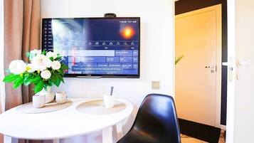 Habitación de hotel con un televisor de pantalla plana montado en la pared mostrando una guía de televisión. Hay una mesa de comedor con un jarrón de flores y algunas tazas. Una silla negra está ubicada frente a la mesa.