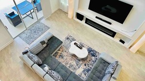 A modern living room with a large flat-screen TV, a grey sectional sofa, a coffee table, and a dining area with blue chairs.