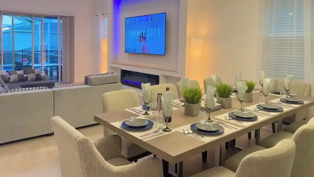 A modern dining area with a long table set for six, a flat-screen TV on the wall, and a large window overlooking a pool.