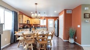 Un comedor amplio con una mesa y sillas de madera, un candelabro y vista a la cocina al fondo.