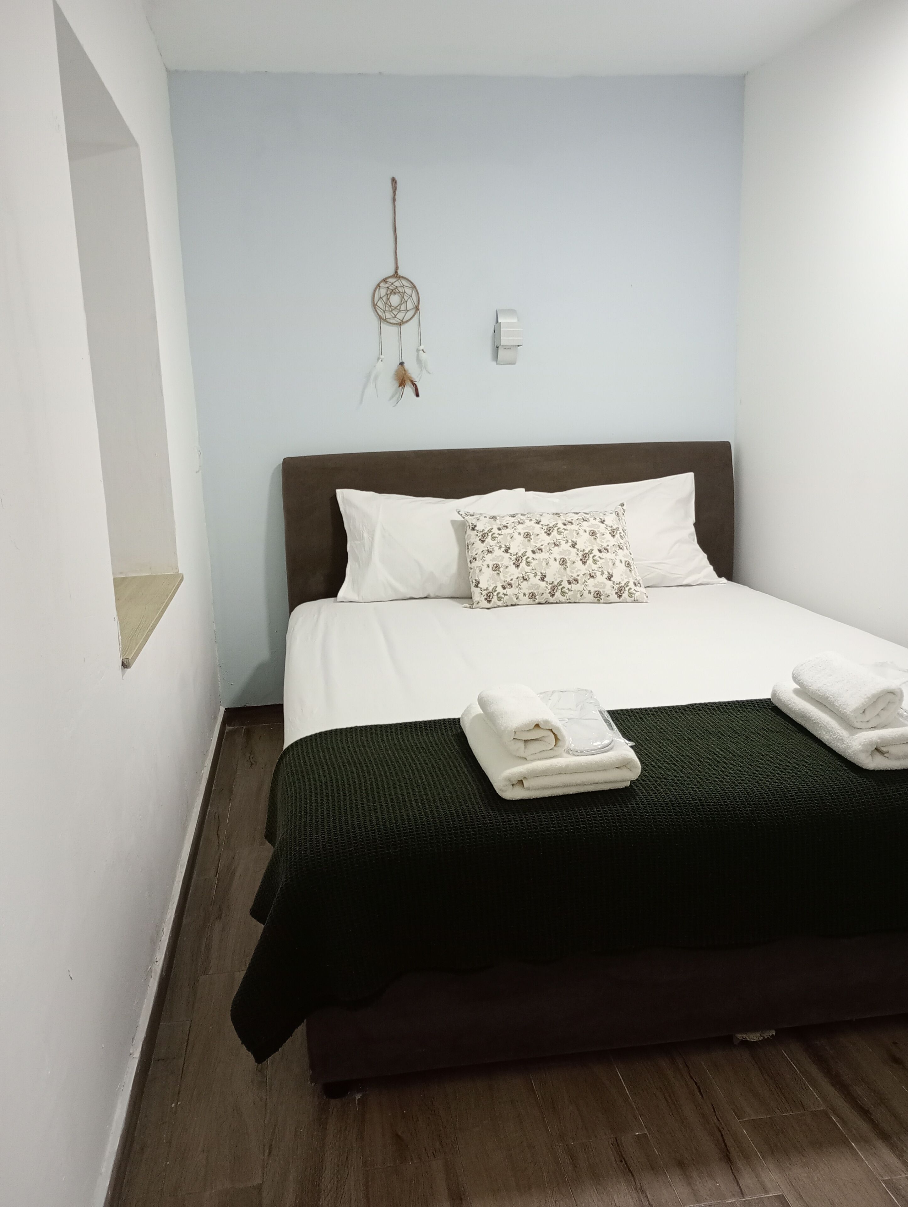A bedroom with a bed, pillows, a dreamcatcher, a wall-mounted light, and a small shelf.