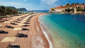 A beach with sun loungers and umbrellas, a clear blue sea, and a coastal town in the background. - SERENİTA STONE HOUSE (Rafajlovici)