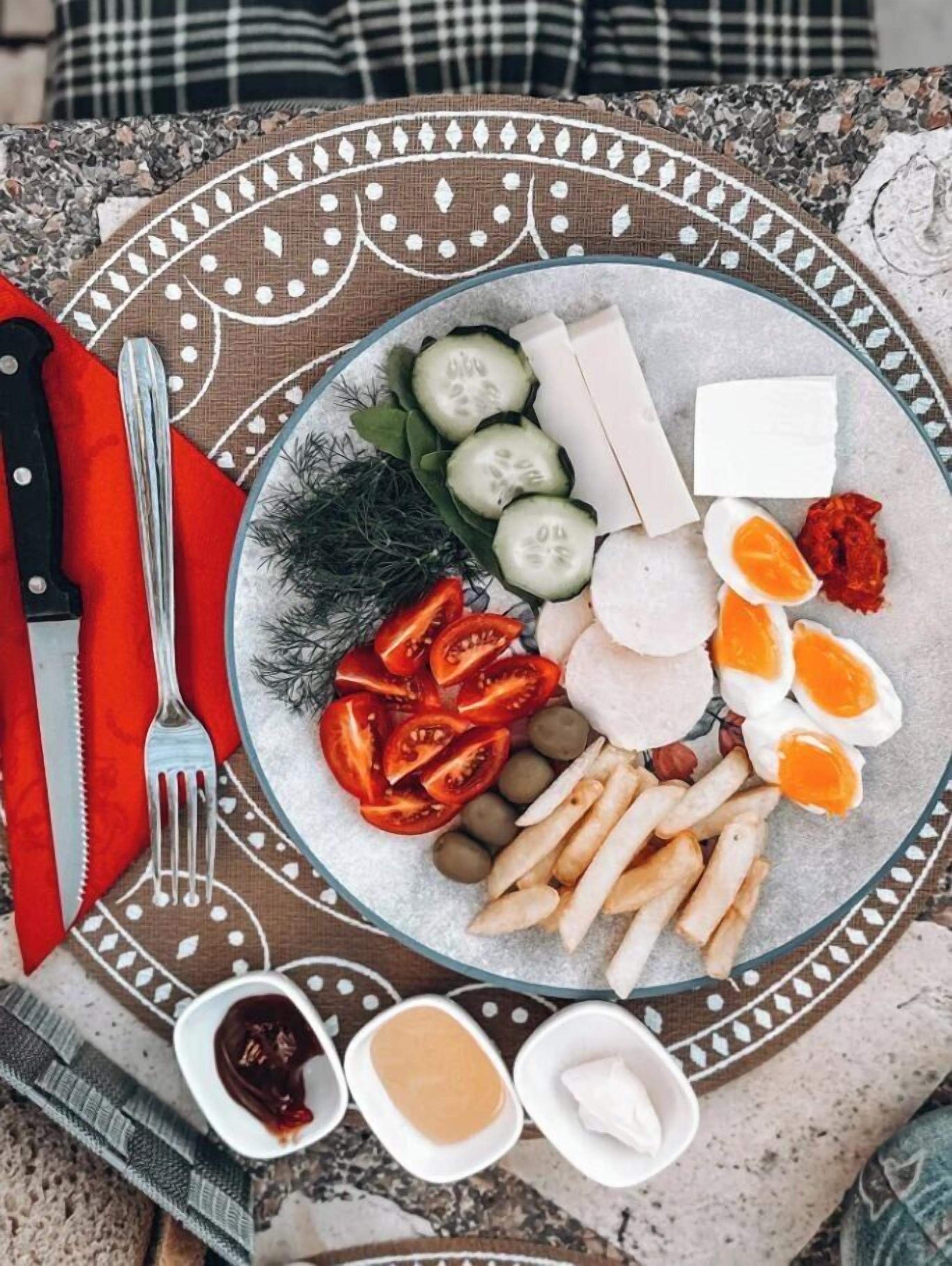 Een bord met diverse soorten eten, zoals gesneden komkommers, tomaten, olijven en eieren, vergezeld van toevoegingen en een mes en vork op een tafelkleed met dessin.