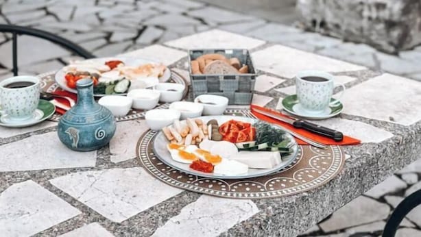 An outdoor dining area with a stone archway, a tiled floor, and a table set with various dishes, cups, and a decorative vase. - SERENİTA STONE HOUSE (Rafajlovici)