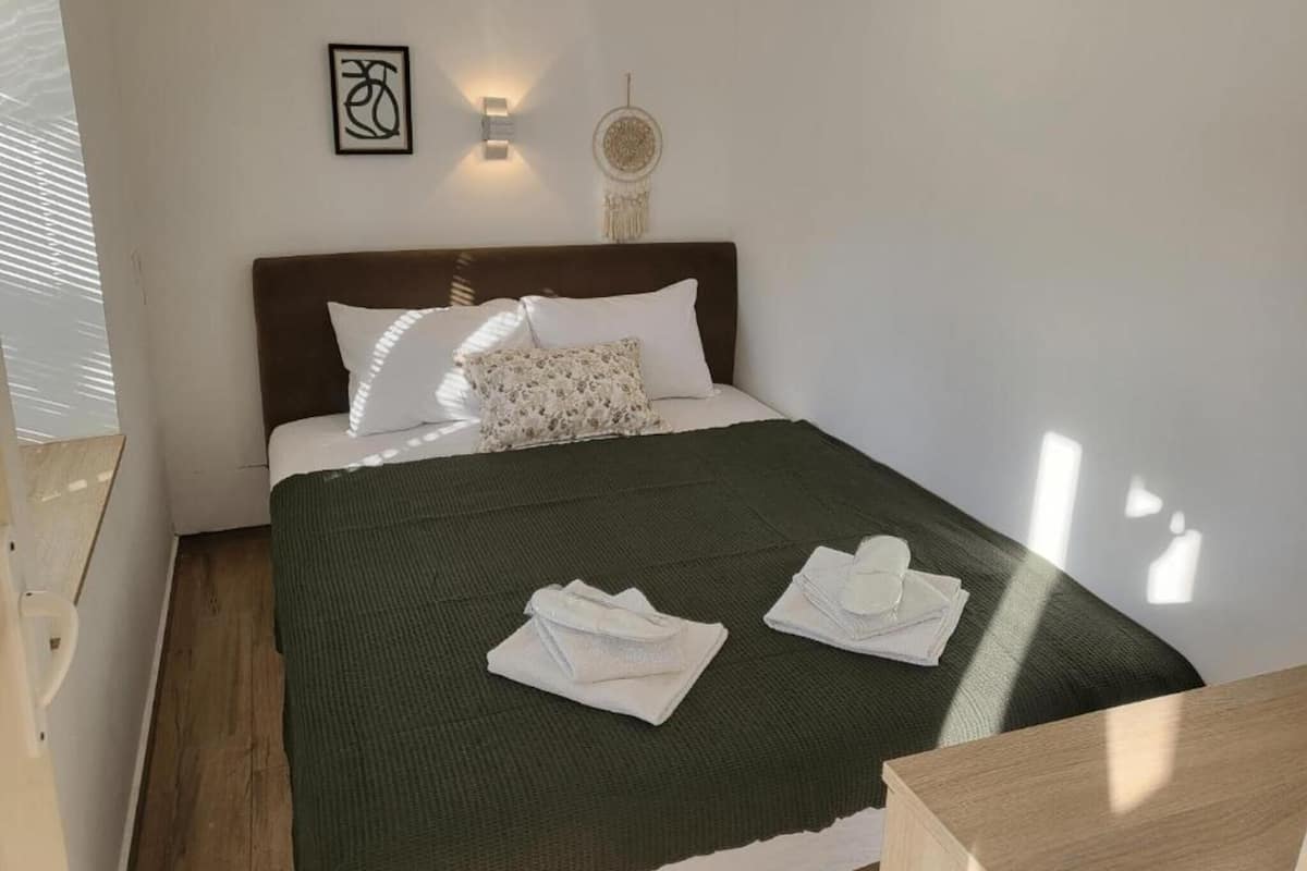 A neatly made bed with a dark green blanket, white pillows, and a headboard. There are two folded white towels on the bed.