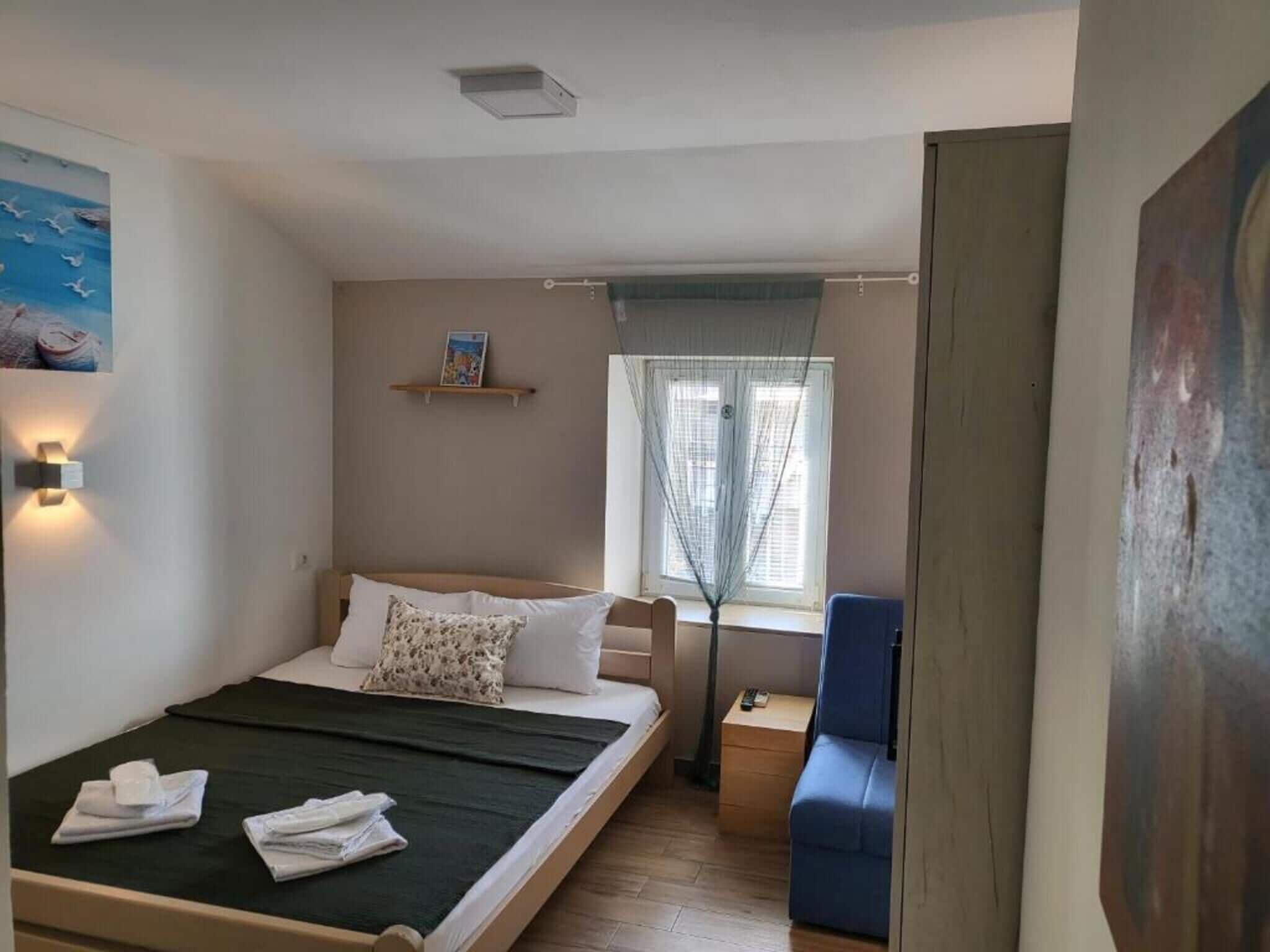 Un dormitorio con cama, mesita de noche, una silla, ventana con persianas y estante en la pared.