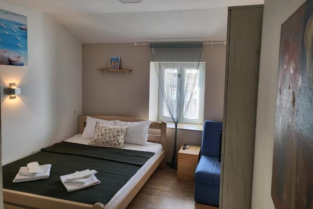 A bedroom with a bed, a nightstand, a chair, a window with blinds, and a wall-mounted shelf.