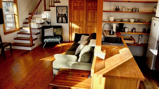 A cozy living room with a wooden staircase, a sofa, and a kitchen area with shelves. - Peaceful private location, cottage @Acadia NP, Lighthouse, Beaches, Seafood🦞 (Bass Harbor)