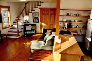 A cozy living room with a wooden staircase, a sofa, and a kitchen area with shelves. - Peaceful private location, cottage @Acadia NP, Lighthouse, Beaches, Seafood🦞 (Bass Harbor)