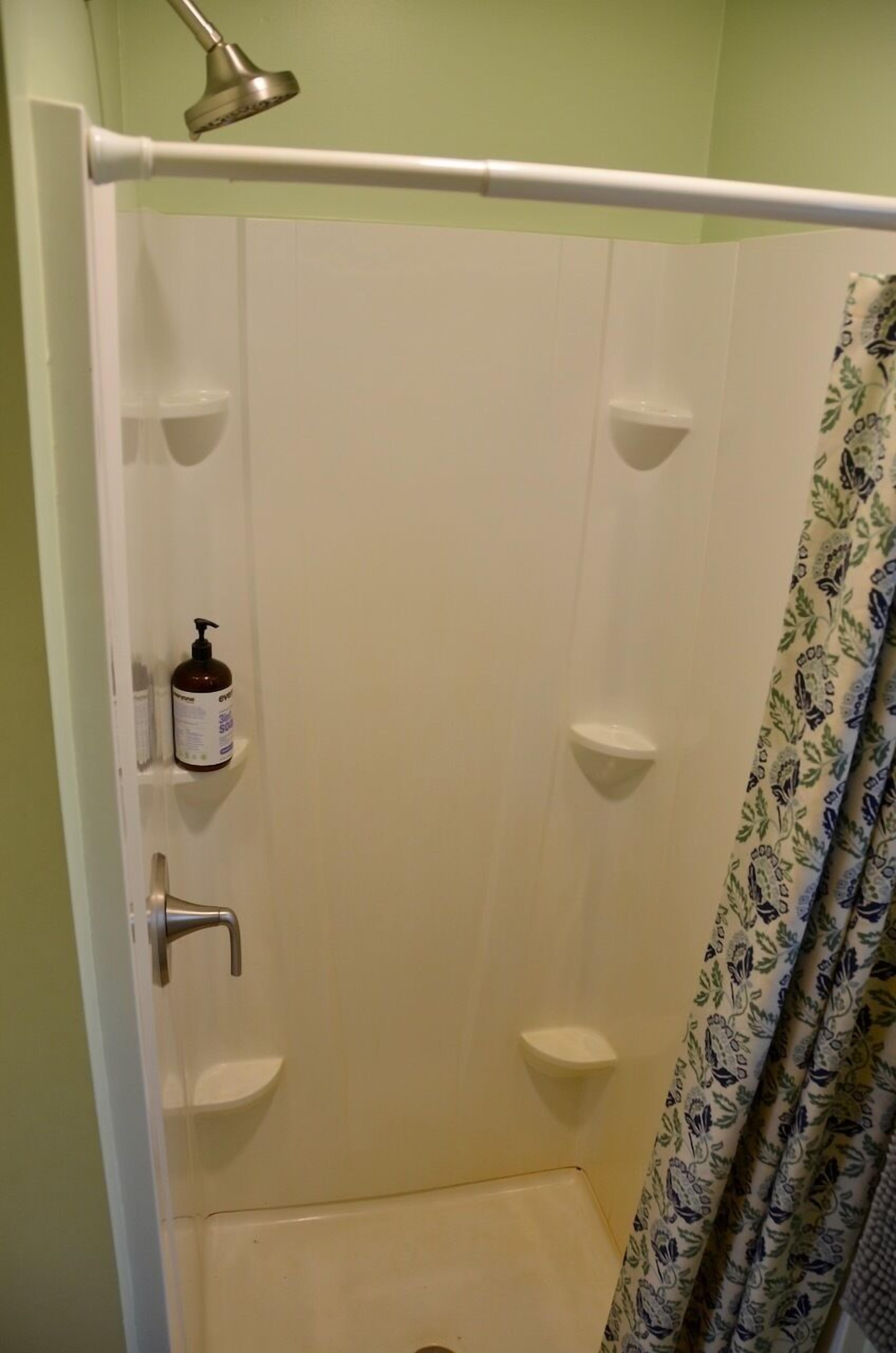 A shower with a white curtain, a handheld showerhead, and a bottle of shampoo on the wall.