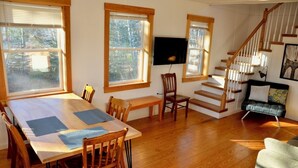 A dining area with a wooden table and chairs, a television mounted on the wall, and a staircase leading to an upper level. - Peaceful private location, cottage @Acadia NP, Lighthouse, Beaches, Seafood🦞 (Bass Harbor)