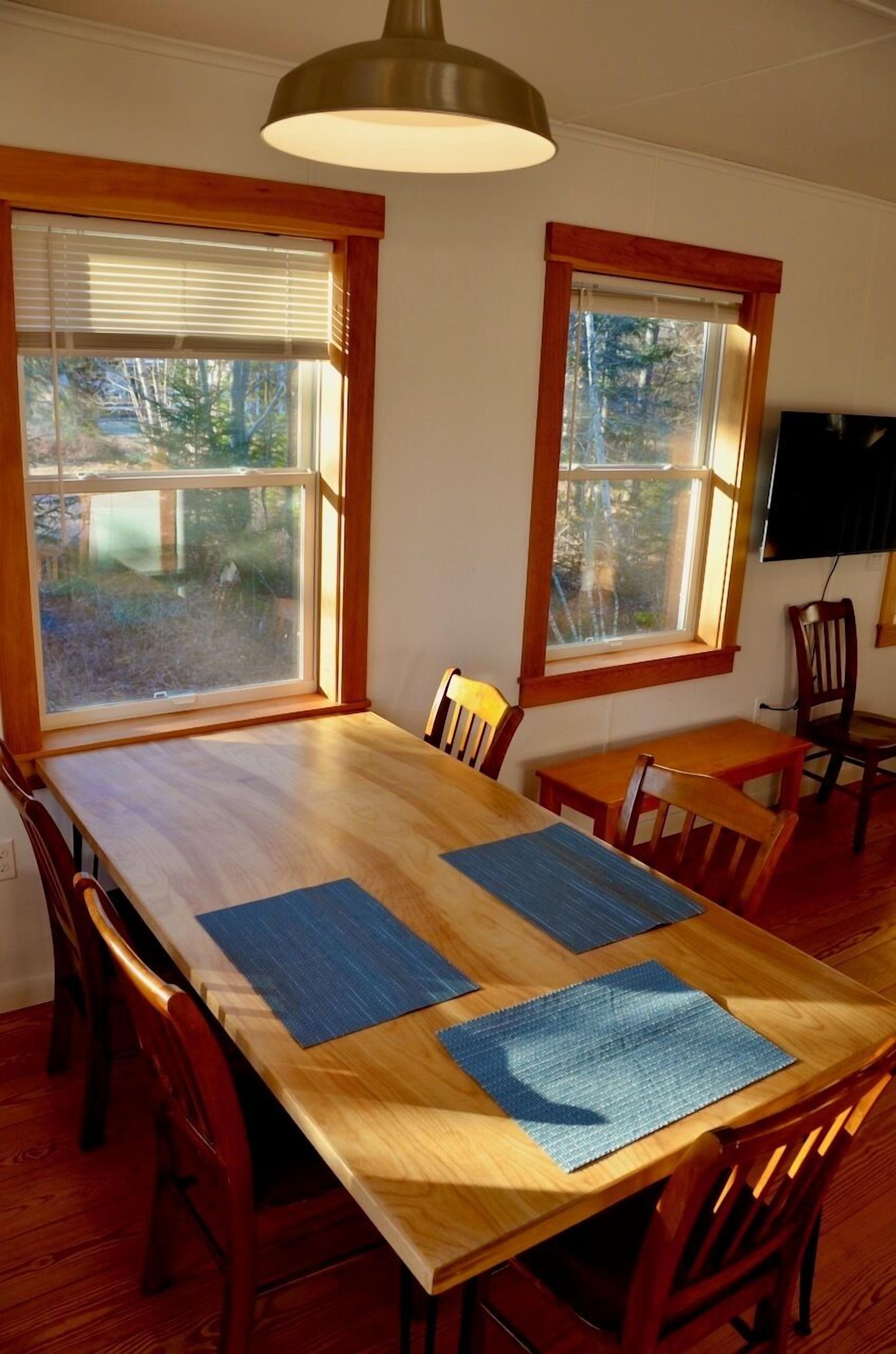 Un comedor con una mesa de madera, cuatro sillas, dos ventanas con persianas y una lámpara colgante.