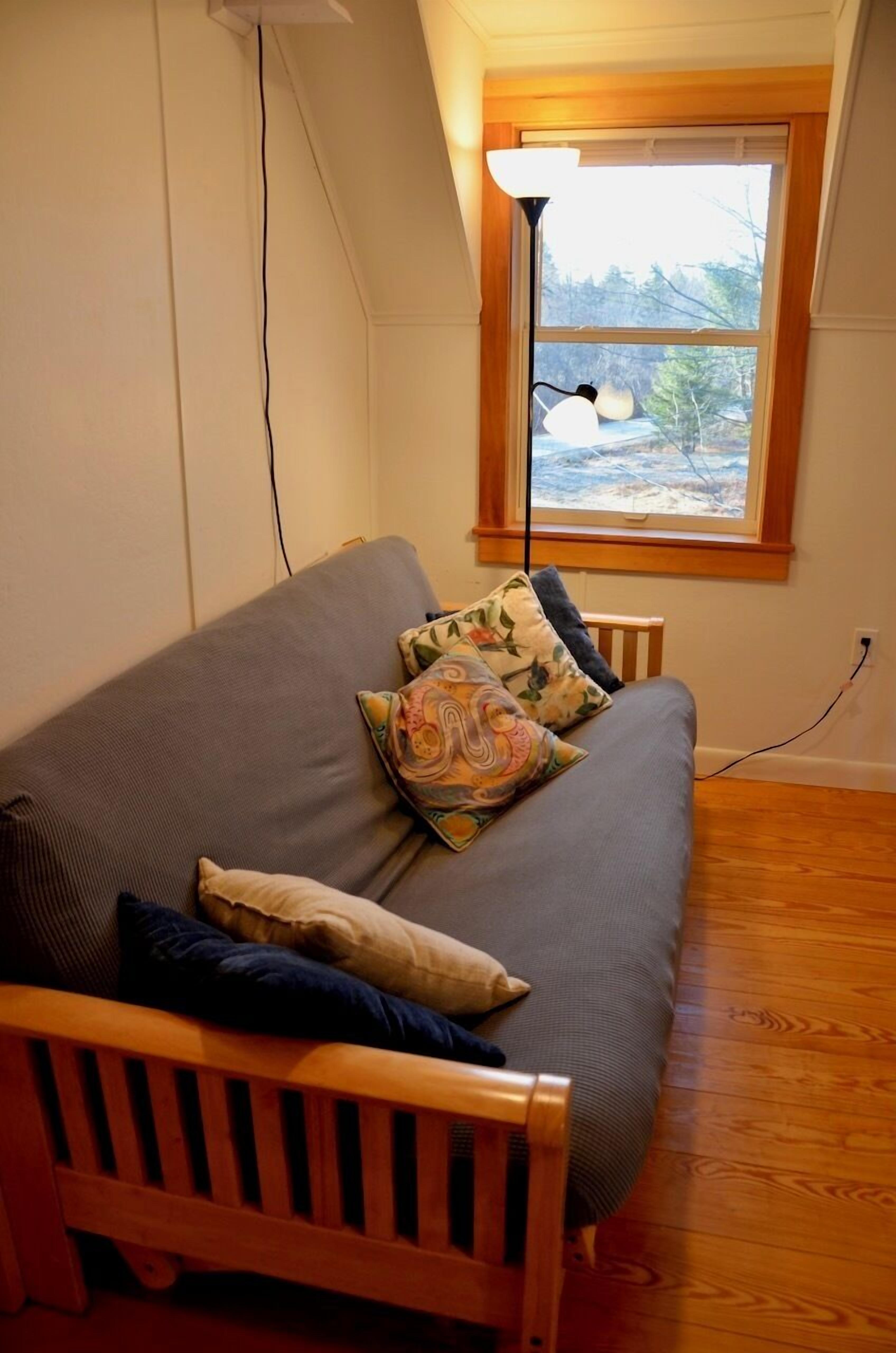 Ein Zimmer mit grauem Sofa, einer Holzbank, einer Lampe und einem Fenster mit Blick auf Bäume.