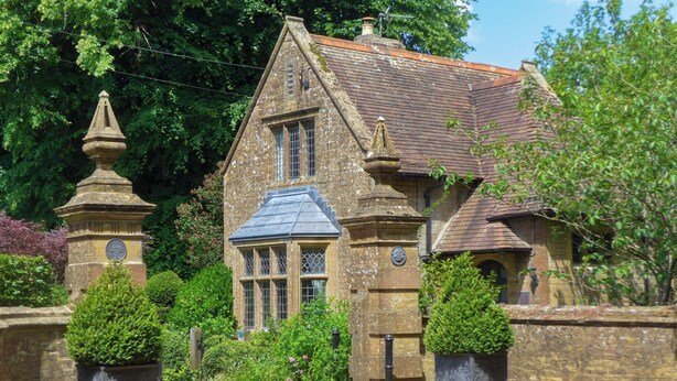 A traditional stone house with a steeply pitched roof, a central window with multiple panes, and two stone pillars with decorative tops. - 2 bedroom accommodation in Crewkerne (Crewkerne)
