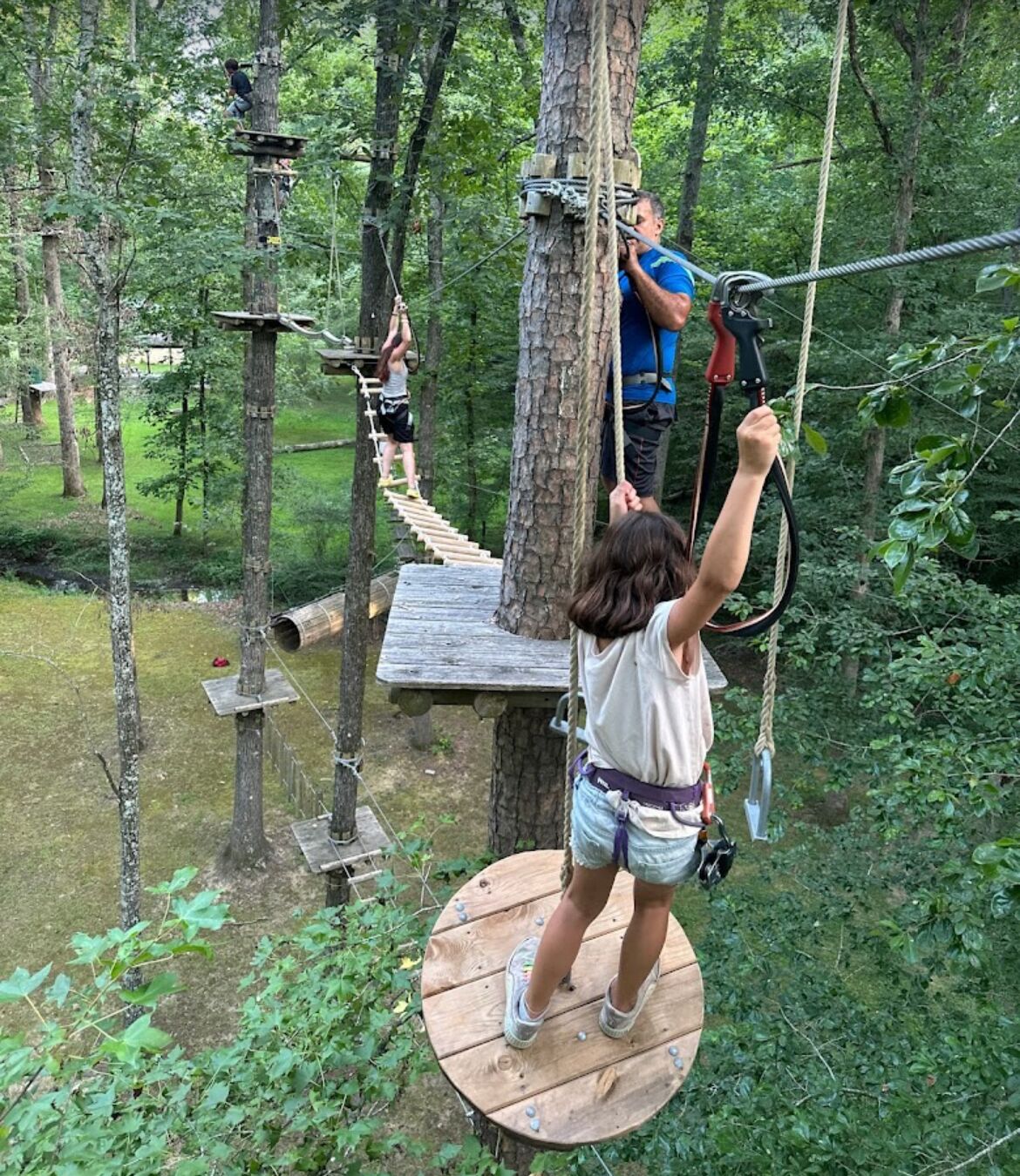 A person on a wooden platform in a forested area with rope bridges and wooden structures.