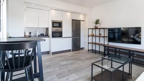 Une cuisine moderne avec des armoires blanches, un four encastré, un four à micro-ondes et un réfrigérateur. On y trouve un coin repas avec une table et des chaises noires, ainsi qu’un téléviseur fixé au mur.