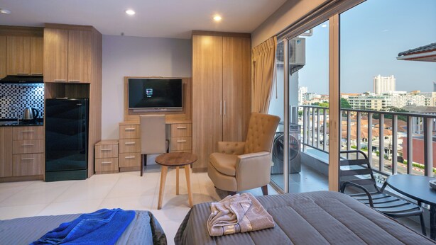 A modern hotel room with a kitchen, a flat-screen TV, a comfortable chair, a small table, and a balcony with a city view. - New Nordic Trend 5 by Apartwell (Pattaya)