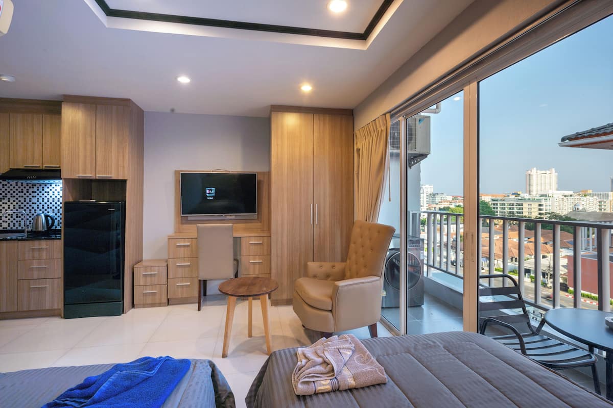 A modern hotel room with a kitchen, a flat-screen TV, a comfortable chair, a small table, and a balcony with a city view.