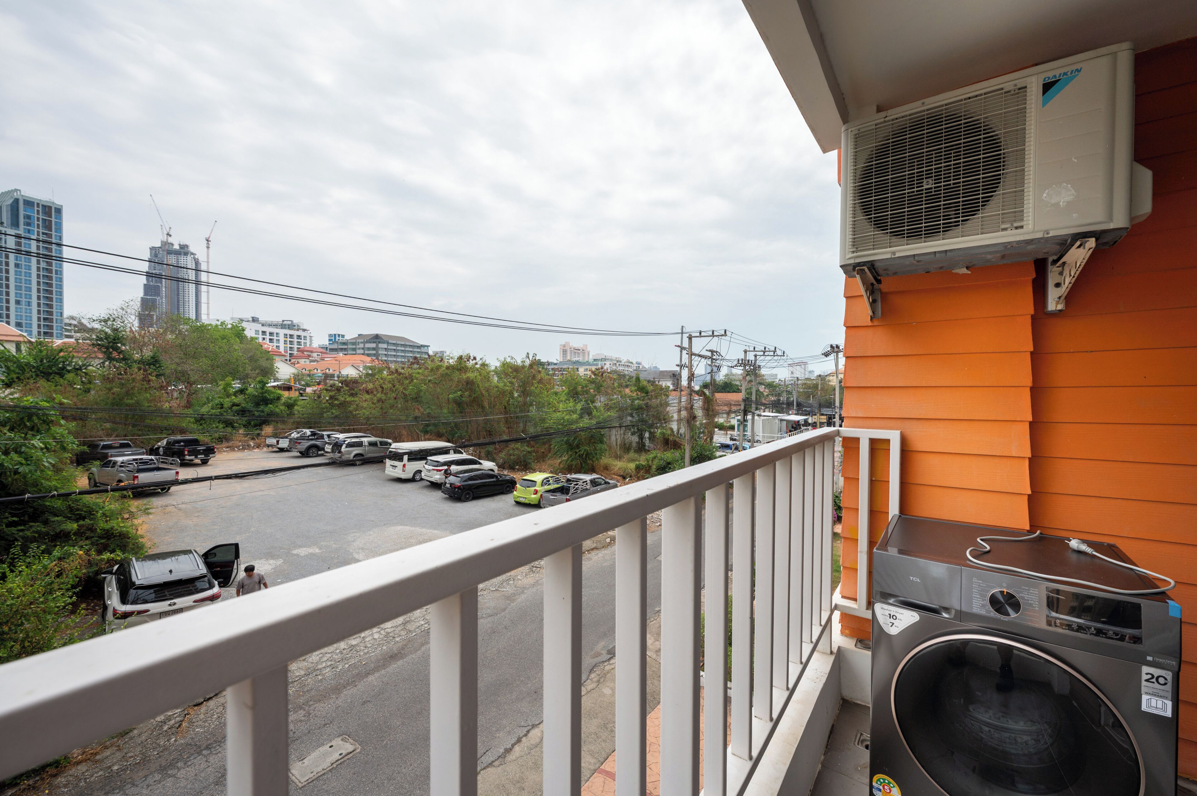 A balcony with an air conditioning unit, a washing machine, and a view of a parking lot with cars and buildings in the background.
