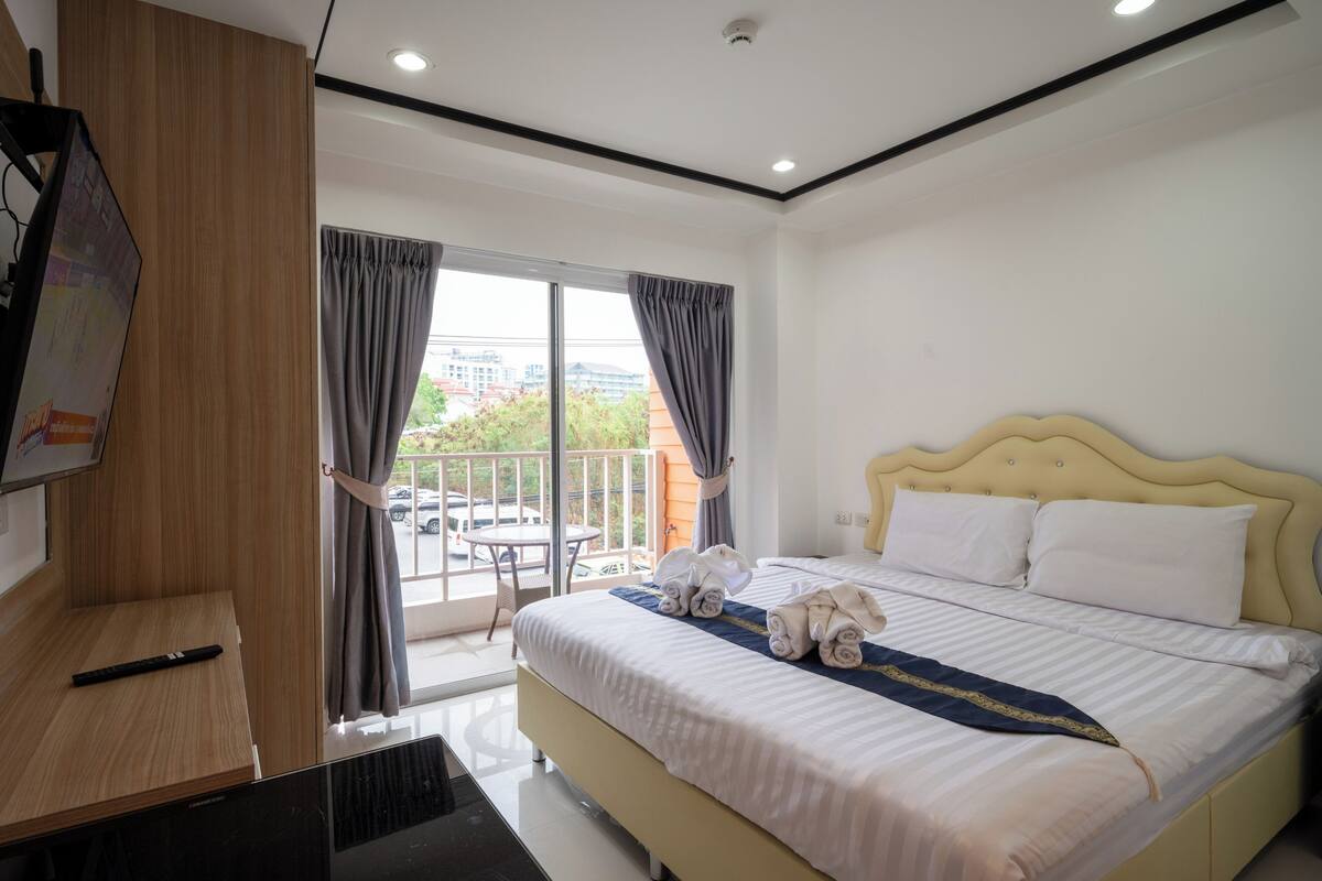 A hotel room with a bed, a TV, a balcony with a table, and a view of buildings outside.