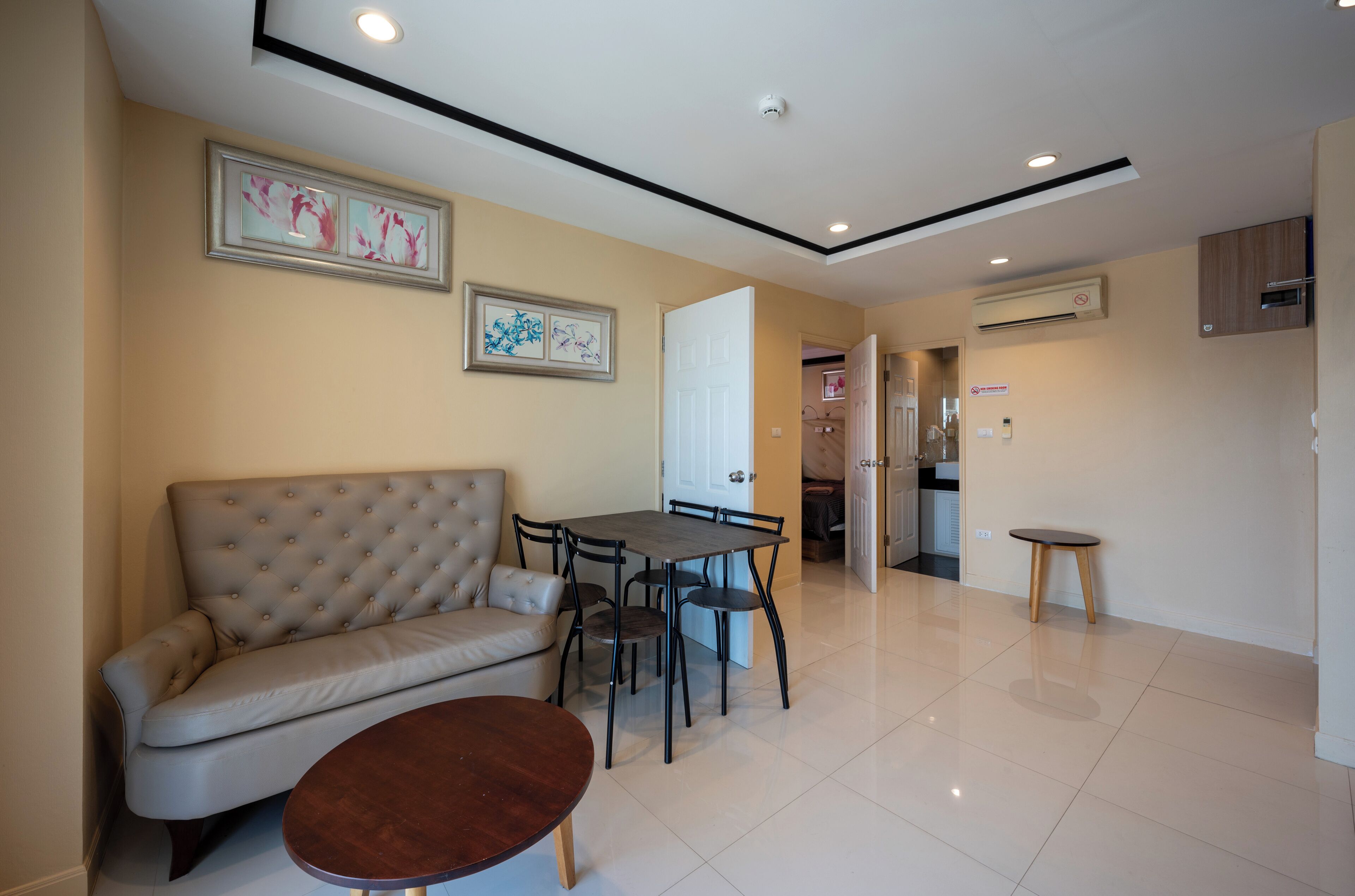 A modern living room with a sofa, dining table, and chairs. There is a small round table in the foreground. The room has a tiled floor and a ceiling with recessed lighting.