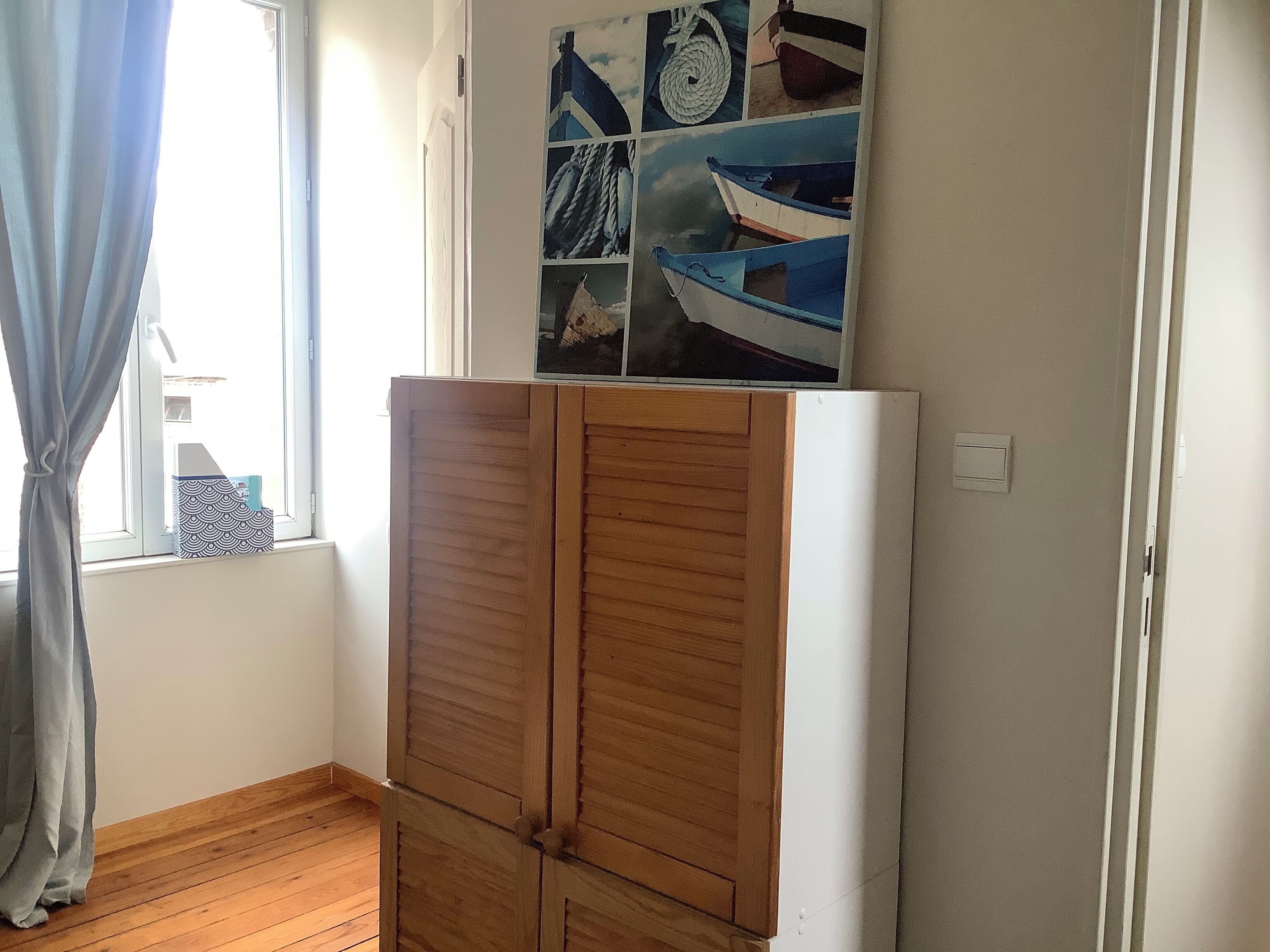 Una habitación con un armario de madera, una ventana con cortinas y una pared decorada con un collage de imágenes de barcos.