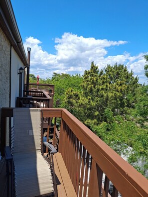 A wooden deck with a railing, a staircase, and a view of trees and a building in the background. - nestled against the dunes, south end beach block on cul-de-sac (Brigantine)