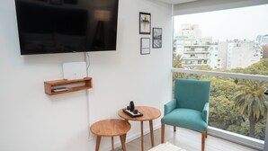 A modern living room with a large TV, a teal armchair, two wooden side tables, and a city view through a large window. - Palermo Cultura Arte y Entretenimiento (Buenos Aires)