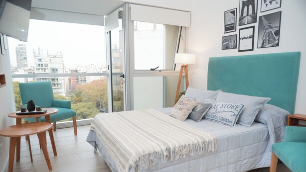 A modern bedroom with a large bed, a teal armchair, a small wooden table, and a city view through a large window. - Palermo Cultura Arte y Entretenimiento (Buenos Aires)