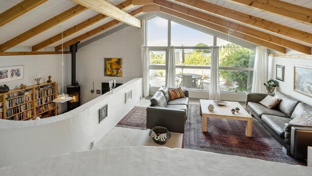 A spacious loft with a high ceiling, exposed wooden beams, a fireplace, a large window, and a cozy seating area with a sofa and a coffee table. - 3 bedroom beautiful home in Kalundborg (Kalundborg)