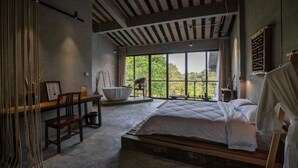 A modern bedroom with a large bed, a wooden desk, and a view of greenery through large windows.