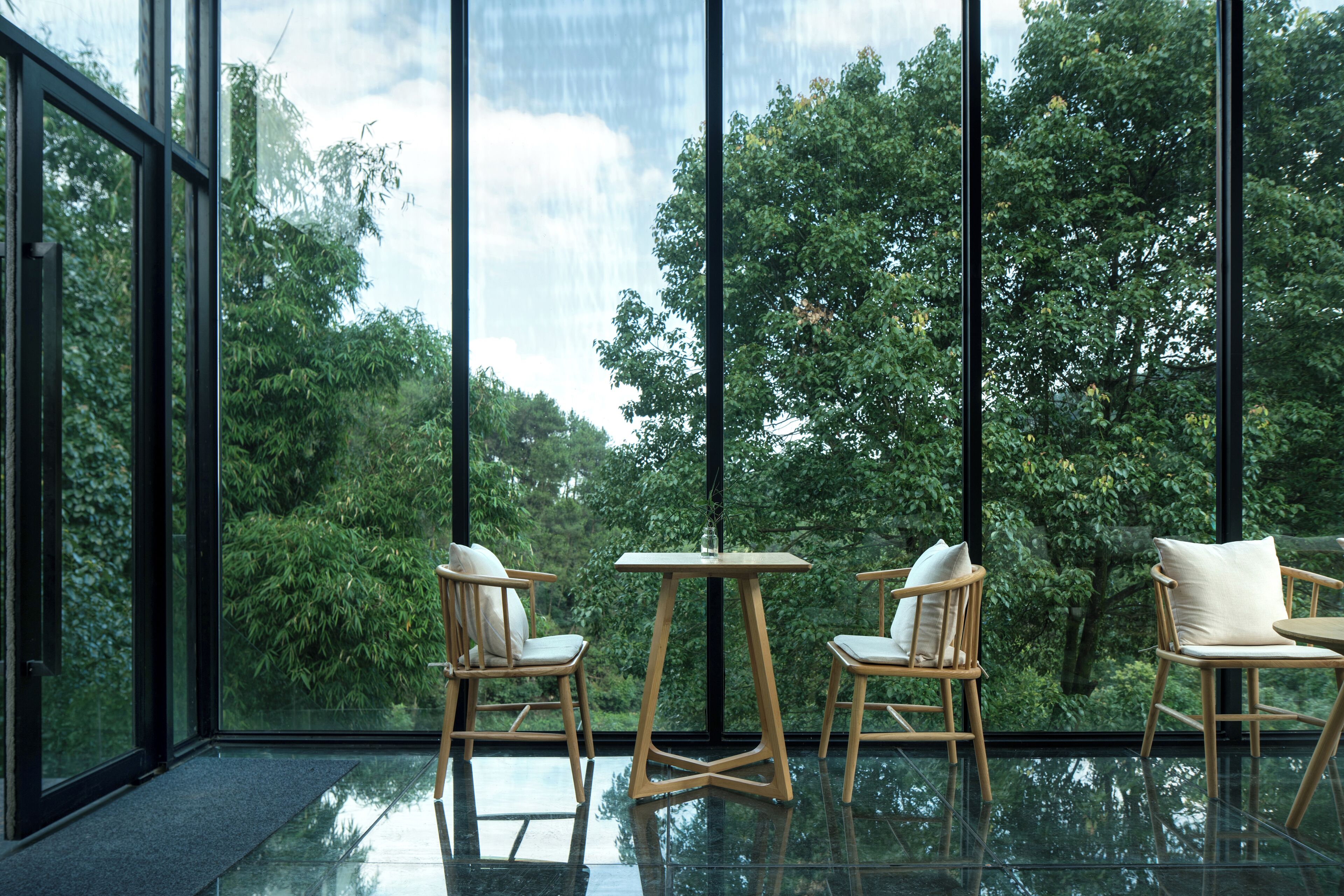 A modern interior with large windows, wooden furniture, and a glass floor reflecting the view of lush greenery outside.