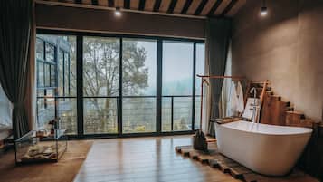 A spacious bathroom with a large bathtub, wooden flooring, and a view of trees through large windows.