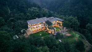 A large, modern house surrounded by dense forest, with multiple levels and large windows.