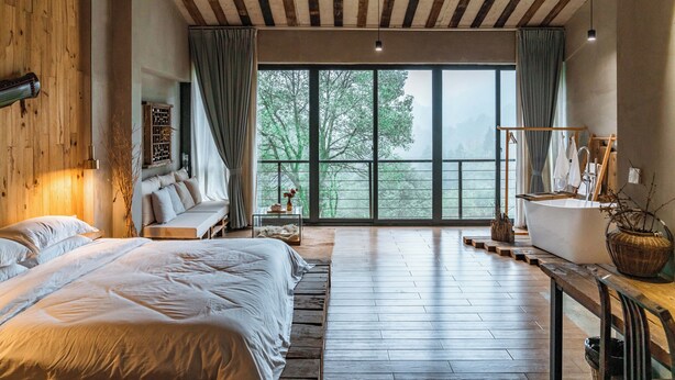 A spacious bedroom with a large bed, a sitting area, a bathtub, and a view of trees through large windows.
