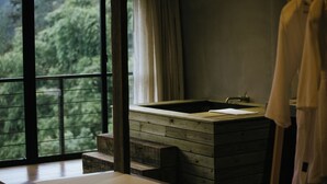 A bathroom with a wooden bathtub, a window with a view of greenery, and a towel hanging on the wall.