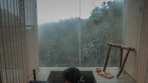 A person in a wooden bathtub with a view of a forest through a mesh window.