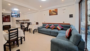 Ein modernes Wohnzimmer mit einem blauen Sofa, einem Essbereich mit Tischen und Stühlen, einem an der Wand befestigten Flachbildfernseher und Kunstwerken an den Wänden.