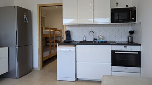 Cocina compacta con refrigerador, microondas, lavavajillas Bosch y horno. Se observa una cama individual en la habitación contigua.