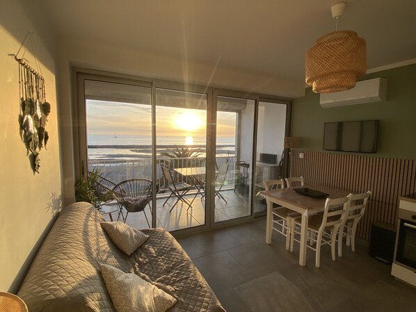 Un balcon offrant une vue sur le coucher de soleil sur la mer, un canapé, une table à manger, des chaises et un téléviseur fixé au mur.