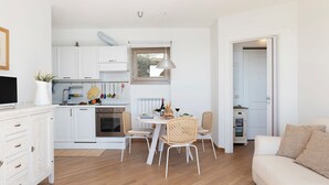 Une cuisine moderne avec des meubles blancs, un coin repas avec des chaises en osier et un canapé blanc dans un espace de vie lumineux et ouvert.