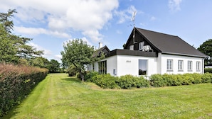 Een modern, twee verdiepingen tellend huis met een witte gevel en donker dak, omgeven door een verzorgd gazon en weelderige struikgewas.