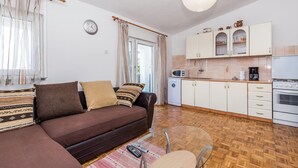 Een compacte woonruimte met een bank, een eettafel, een keukenhoek met kastjes en apparatuur, en een groot raam met gordijnen.