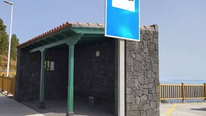 Un arrêt de bus avec un mur en pierre et un toit végétalisé, un panneau indiquant l’arrêt de bus avec un symbole de bus, et une clôture en bois en arrière-plan.