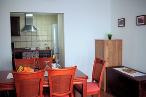 A dining area with a table set for two, chairs, a kitchen in the background, and a cabinet with a plant on top. - Rabel House (La Fajana)