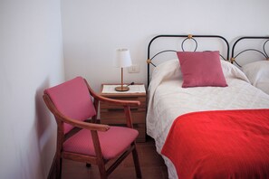 A single bed with a red blanket, a wooden chair with a red cushion, a bedside table with a lamp, and a wall socket.