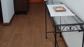 Un couloir avec une porte en bois, une commode sombre, une table en verre ornée d’un objet décoratif et un miroir accroché au mur.