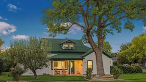 Une maison de plain-pied avec un toit végétalisé, un porche à l’avant doté de deux chaises et un grand arbre devant.
