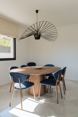Een ronde houten tafel met zes stoelen, een moderne hanglamp en een raam met uitzicht op groen.