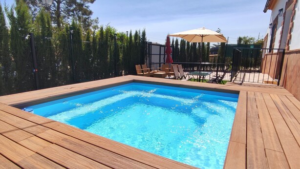 A rectangular swimming pool with wooden deck surrounding it, a patio area with chairs and an umbrella, and a fence with trees in the background.